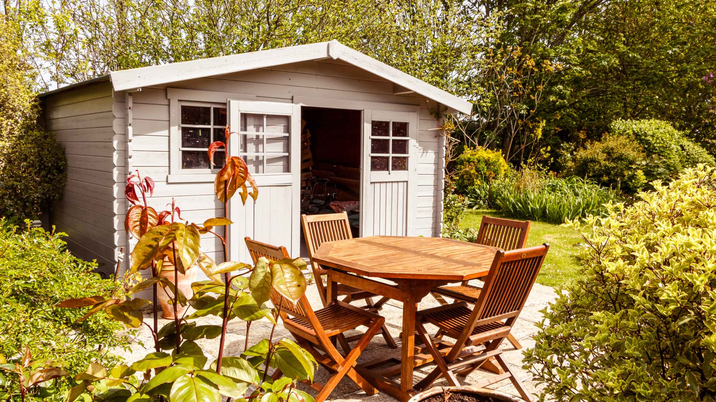 Garden shed surrounded by greenery, it's door is wide open, and there's a table and chairs in front of it.