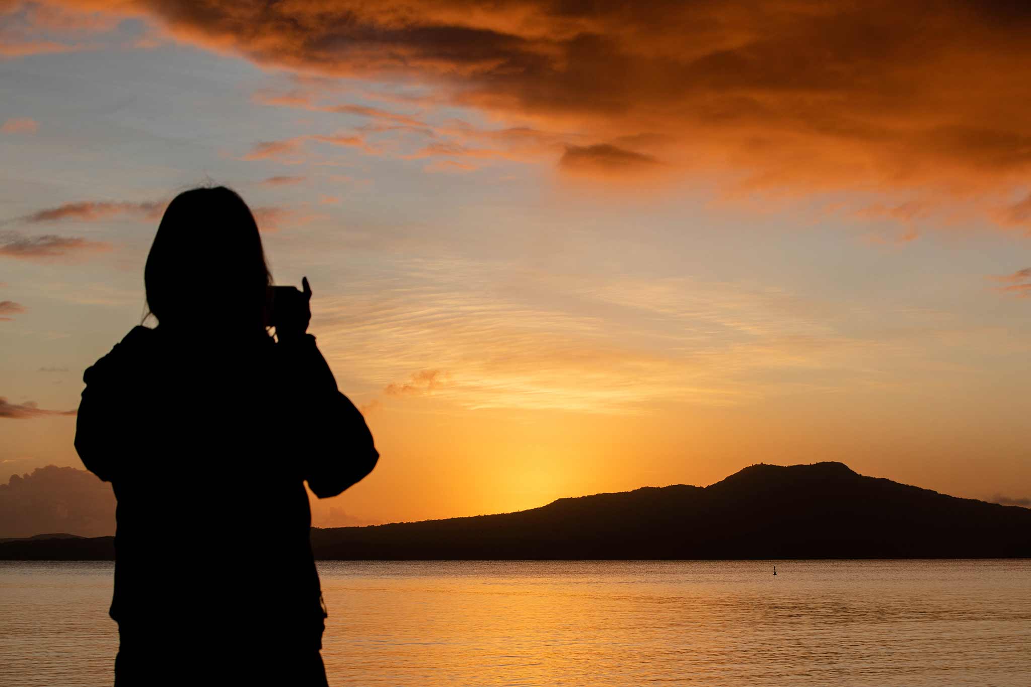 Photo of the silhouette of a person taking a photo of the sunrise over Rangitoto Island.