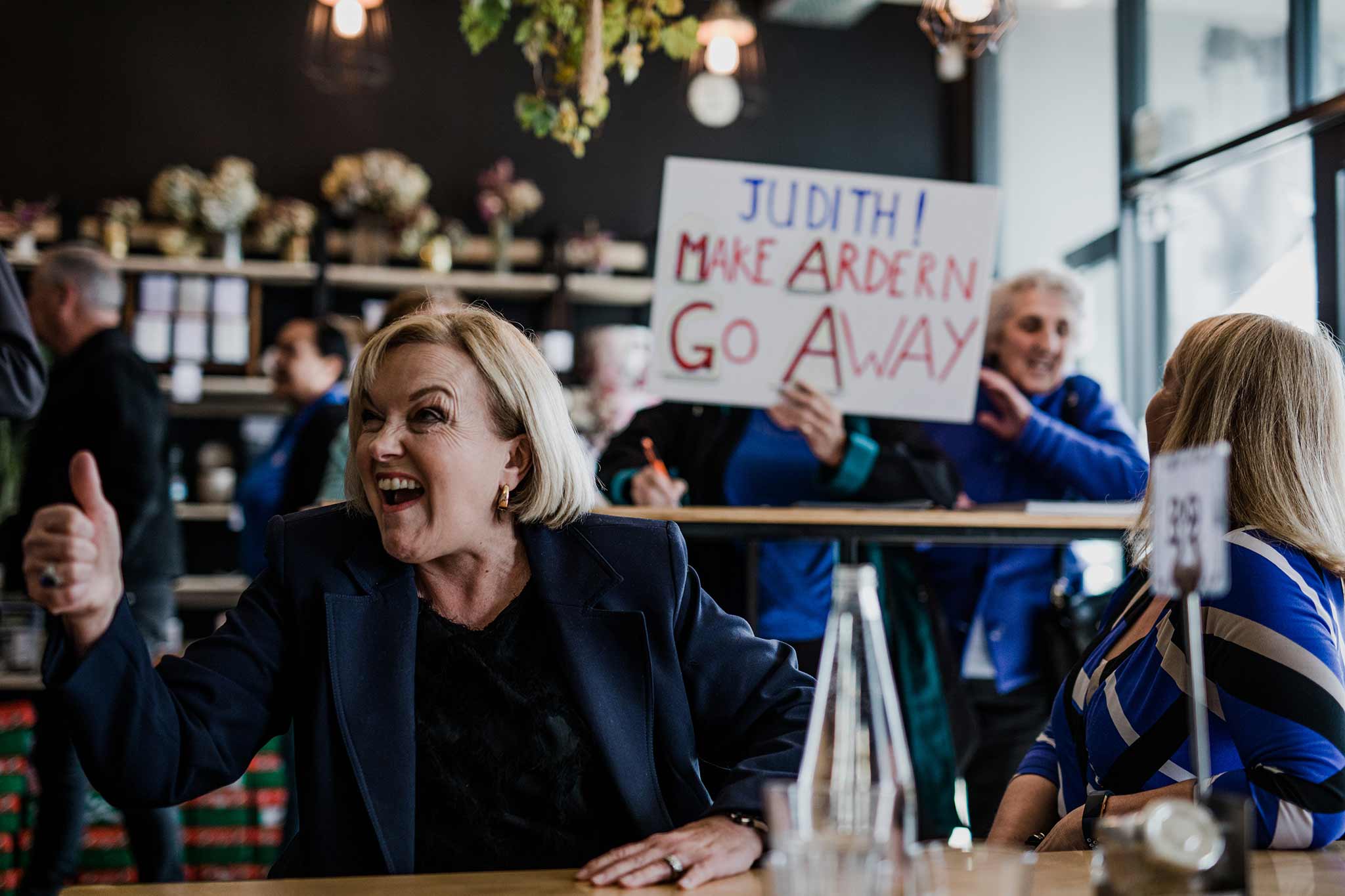 Photo of Judith Collins enthusiastically giving the thumbs up in the foreground, with someone in the background holding a sign saying "Judith! Make Ardern Go Away."