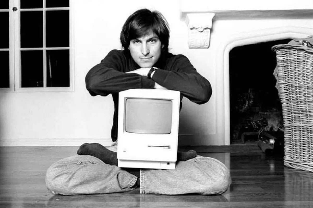 Steeve Jobs posing in front of a camera with one of the first Machintosh