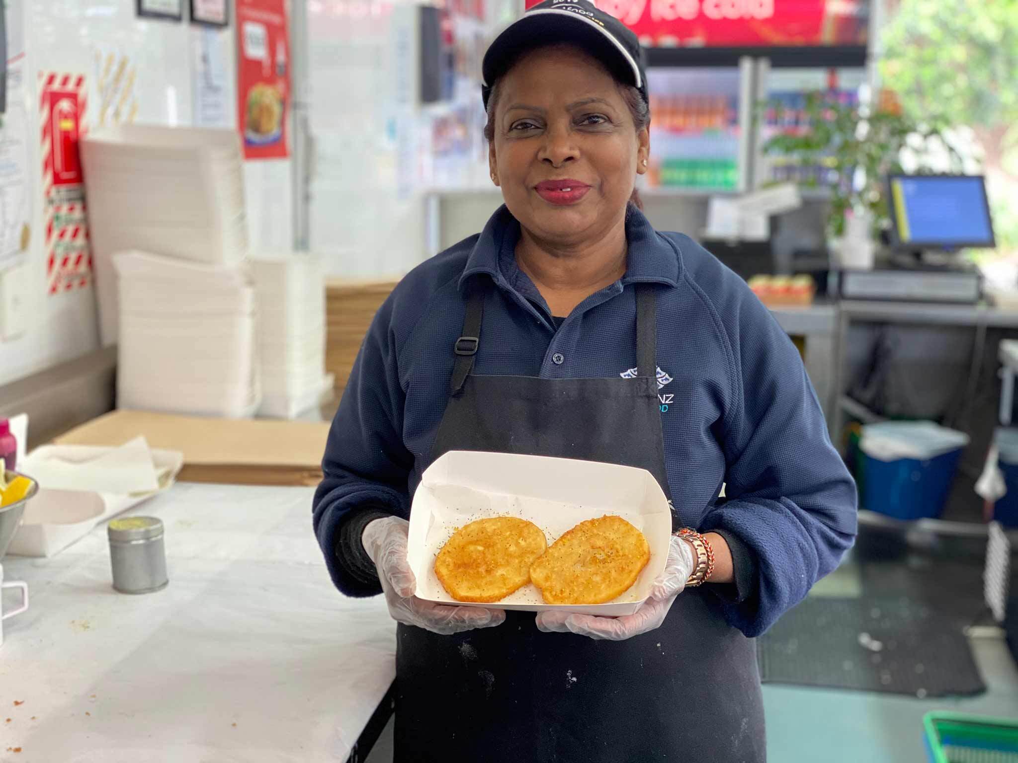 Photo of a woman in a restaurant kitchen holding a cardboard tray with two potato fritters in it.