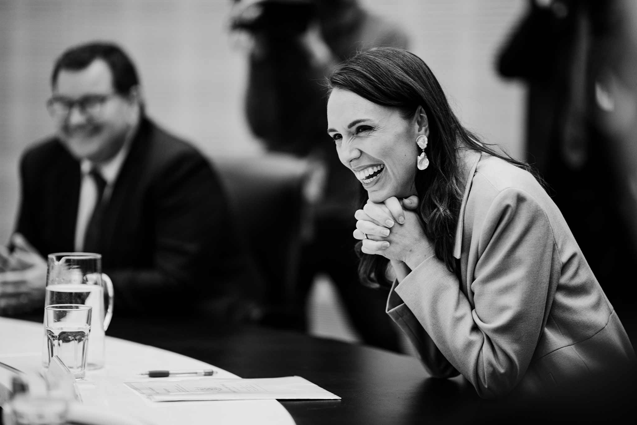 Black and white photo of Jacinda Ardern smiling and leaning on a table, with Grant Robertson smiling in the background.