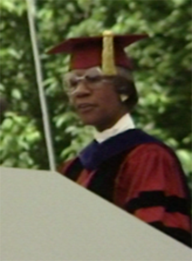 Shirley Chisholm, wearing commencement attire, speaking at podium on outdoor stage, trees in background