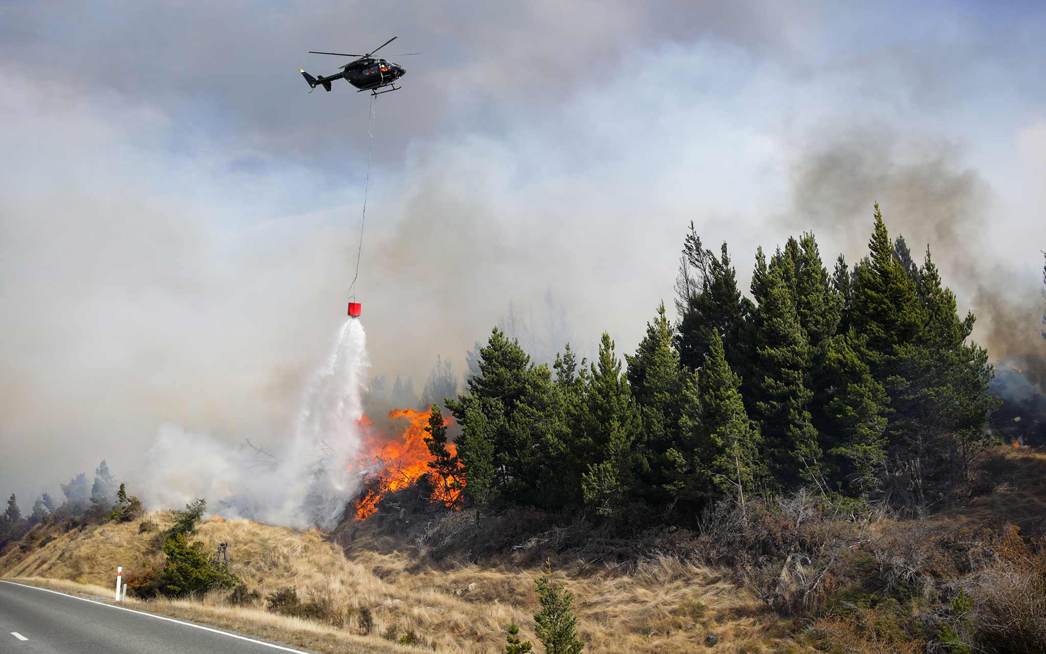 Photo of a helicopter dropping water on a fire in some tress next to a road.