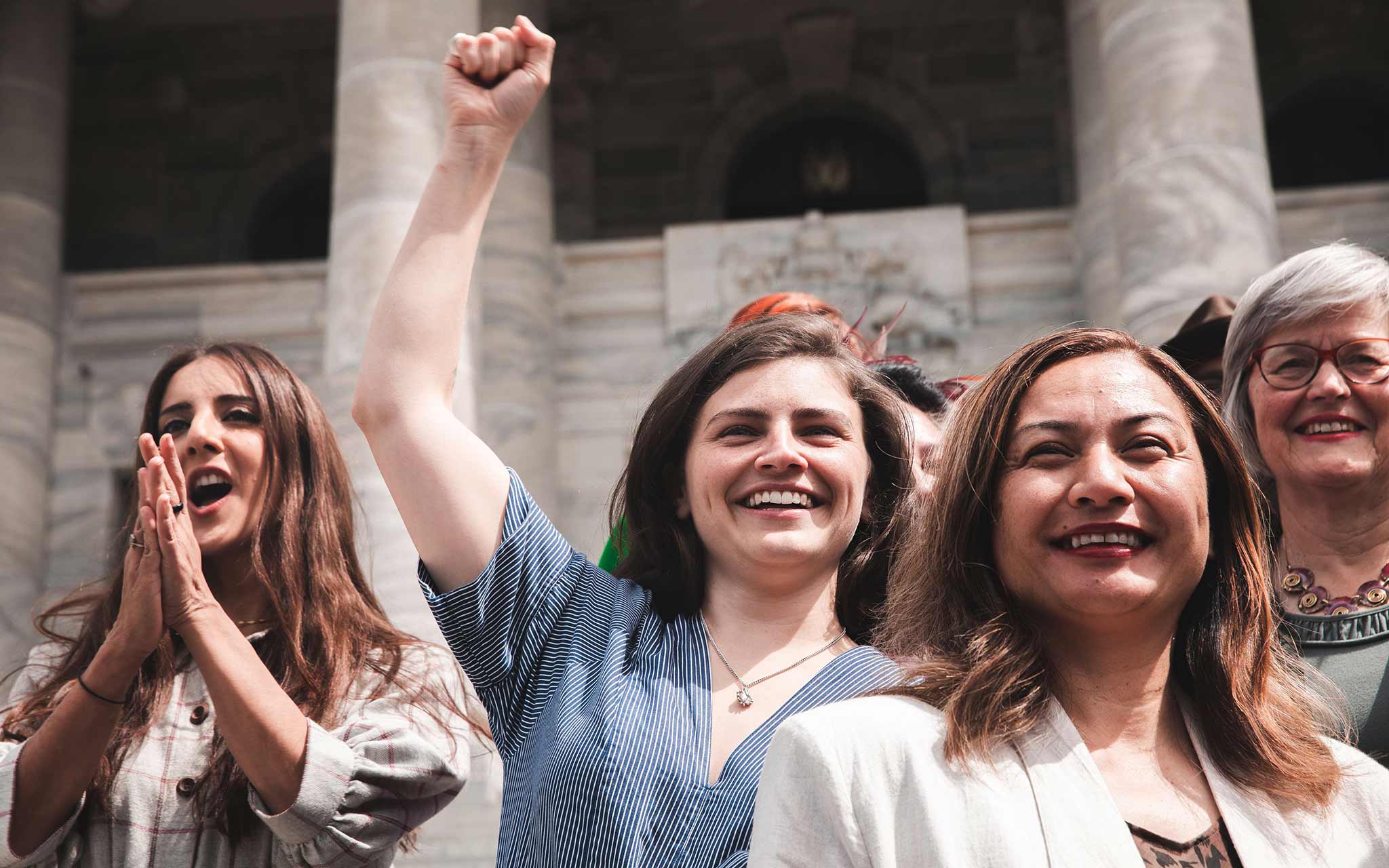 Photo of Golriz Ghahraman, Chloe Swarbrick, Marama Davidson and Eugenie Sage with big smiles, standing in front of Parliament.