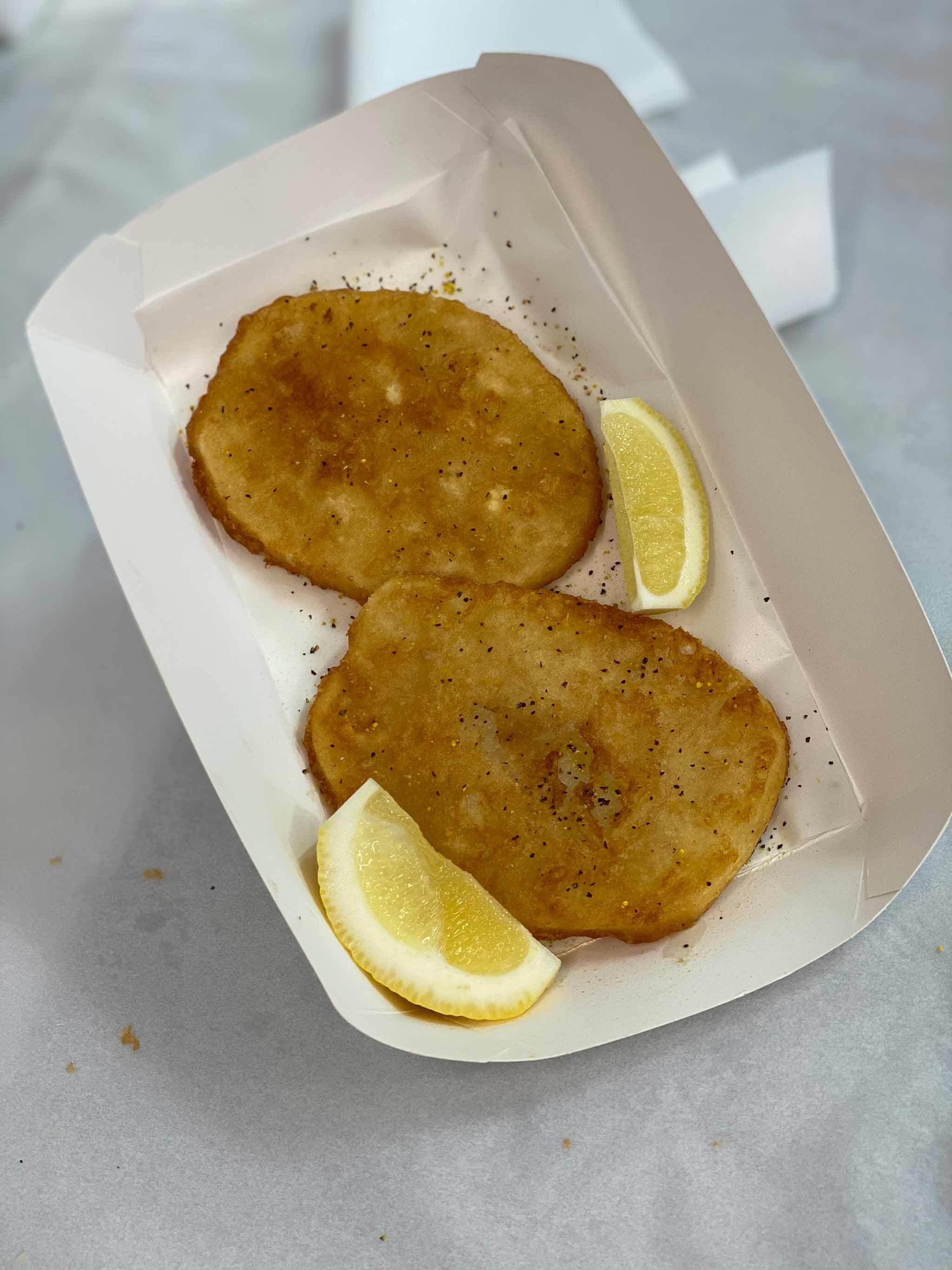 Photo of two potato fritters in a cardboard tray with two slices of lemon.
