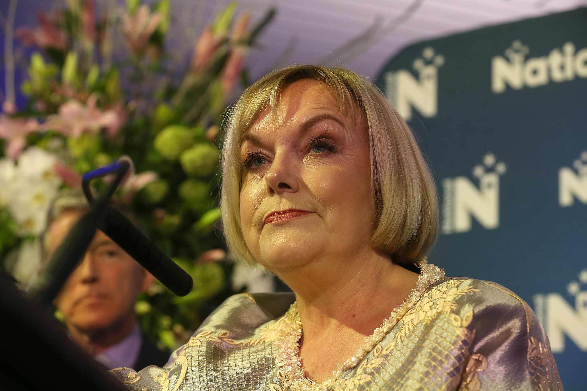 Photo of Judith Collins standing behind a microphone and in front of a National backdrop.