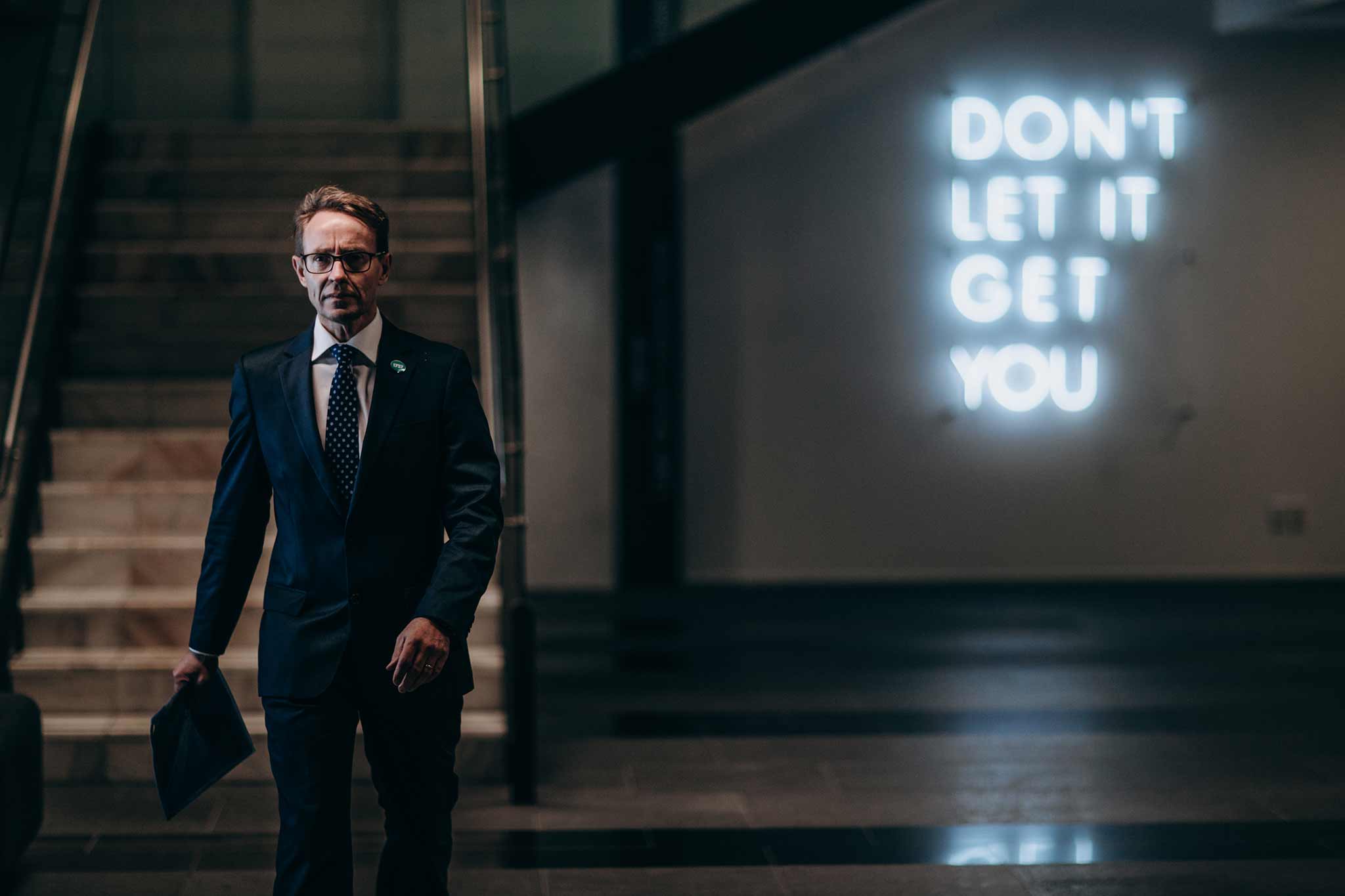 Photo of Ashley Bloomfield walking towards the camera with a neon sign in the background that says, "Don't Let It Get You."