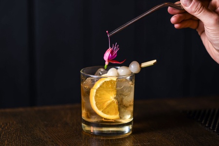 A signature cocktail with ice and a citrus slice, being garnished with a fuschia flower