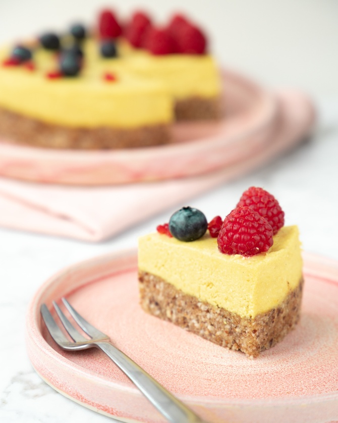 Rød tallerken med et stykke gul raw lemon cheesecake, med hindbær og blåbær på toppen