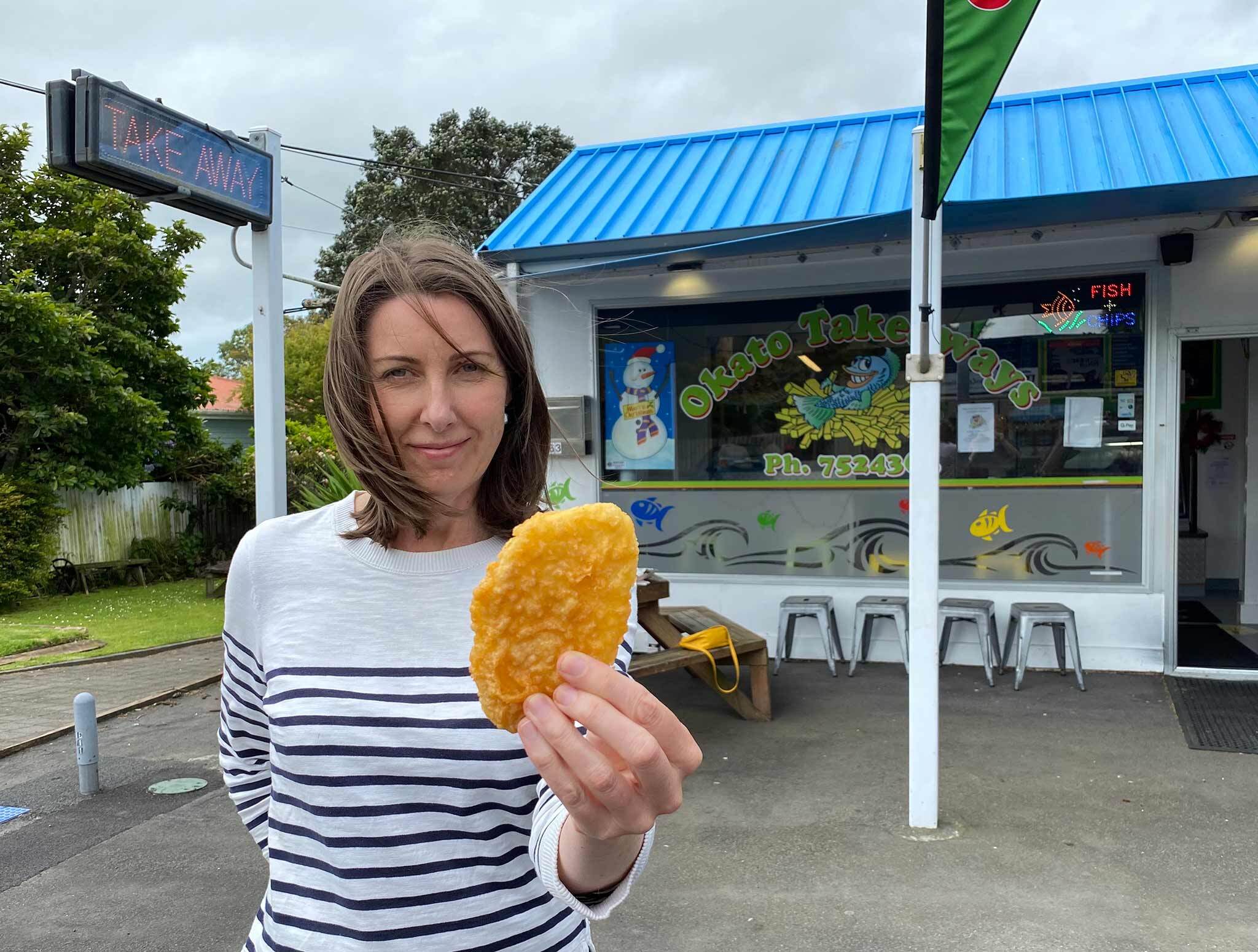 Photo of a woman holding a potato fritter standing in front of Okato Takeaways shop.